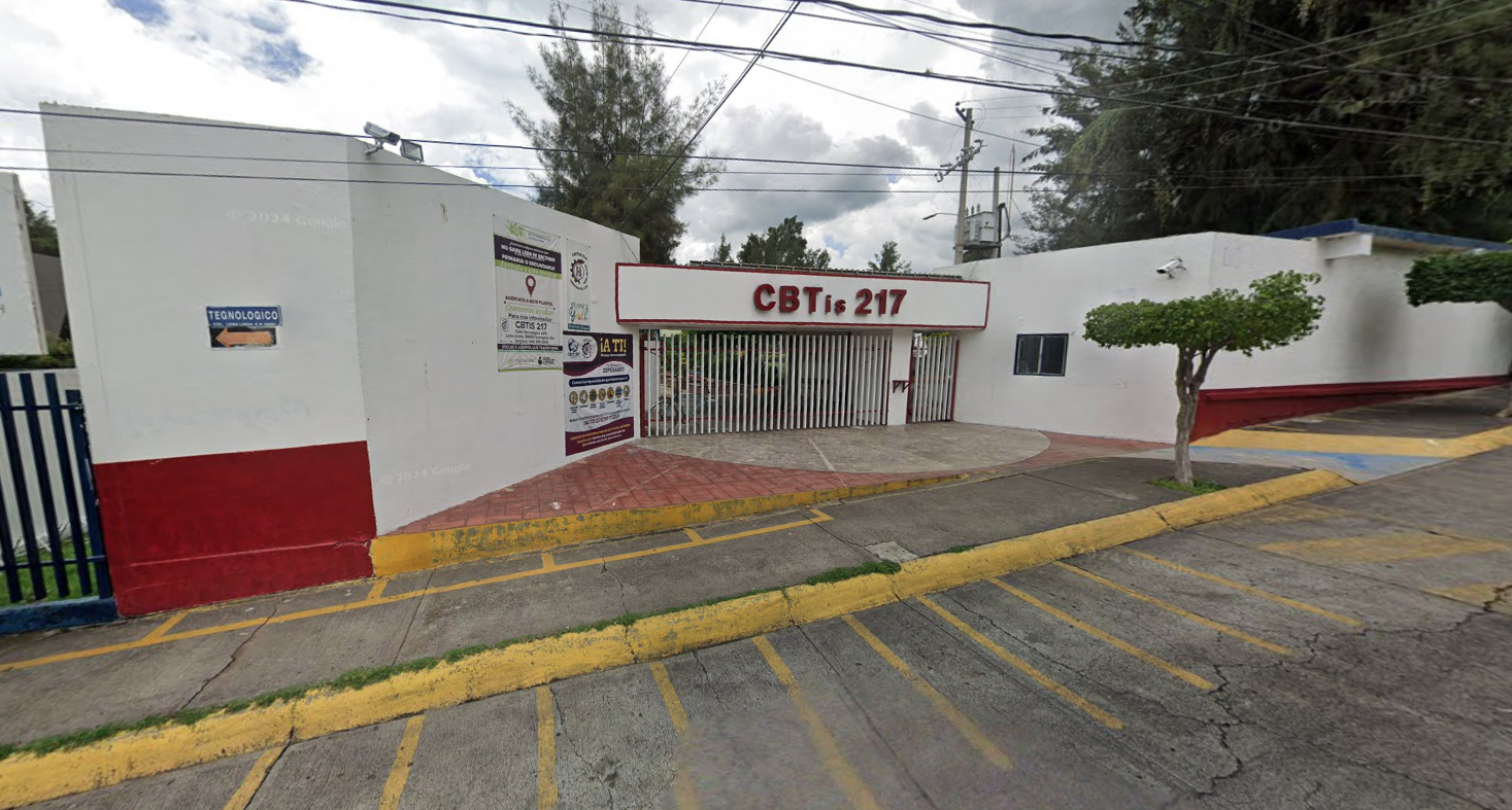Fotografía en la que se muestra la entrada del plantel, conformada por muros de color blanco que rodean las instalaciones. En medio de los muros hay una reja blanca y sobre ell<a hay un muro que en letras rojas grandes dice Cebetis 217.
